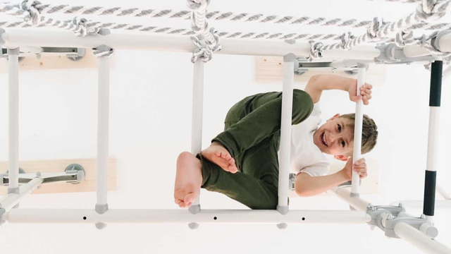 Smiling boy climbing across the overhead monkey bar rungs of a white Brainrich indoor play gym