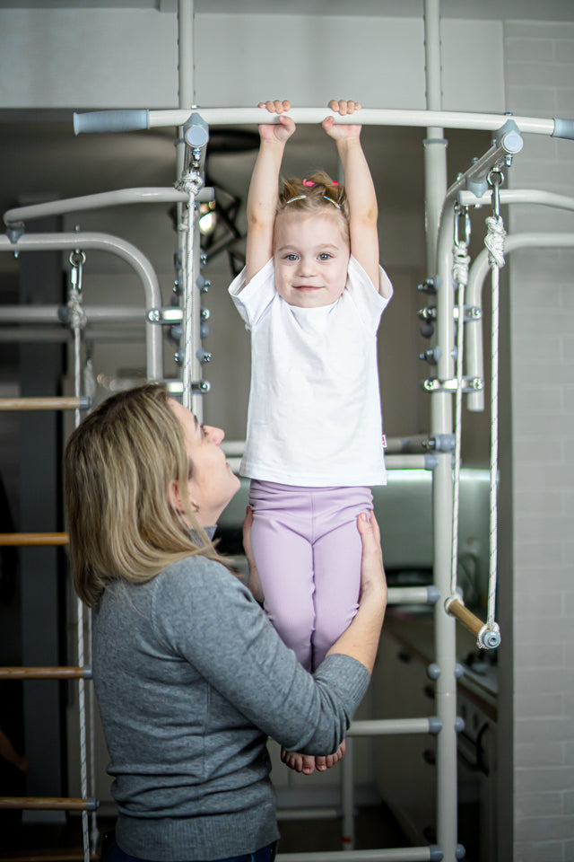 Mother holding her toddler who hangs from an HSA/FSA eligible indoor play gym bar