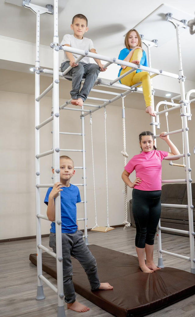 Four kids of different ages playing together on a white Brainrich Mega Max home play gym with crash mats in a living room