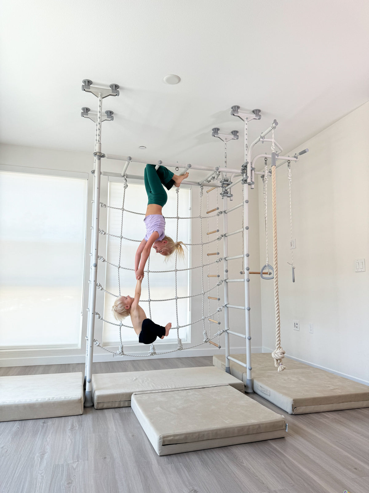 Girl hanging upside down from the bar of a Brainrich play gym while holding her younger brother's hands