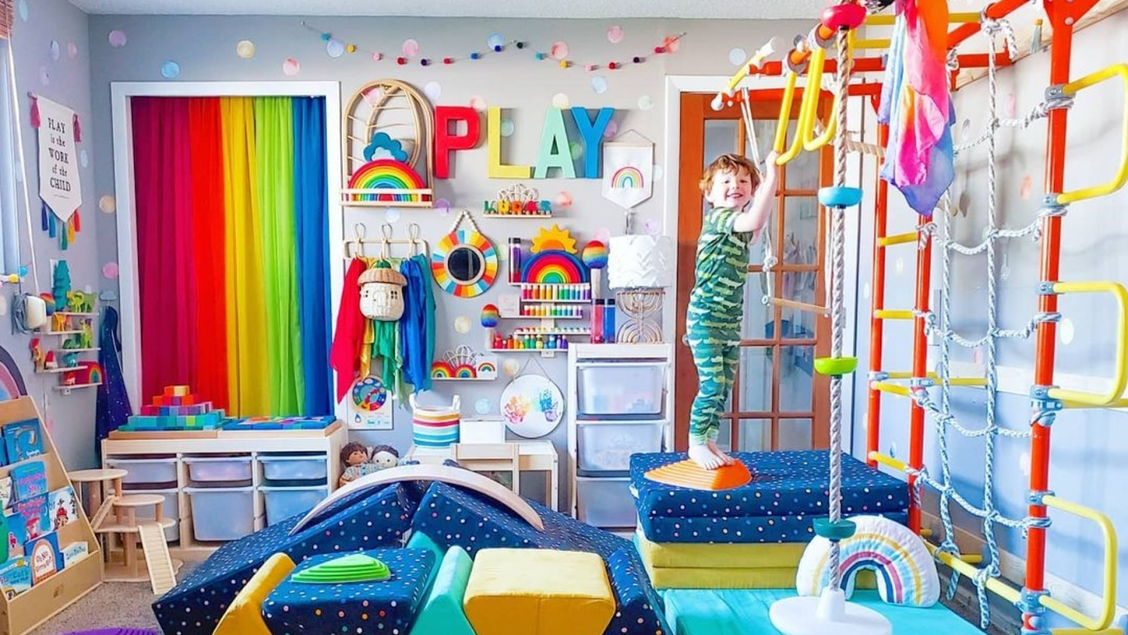 A colorful playroom-homeschool combo room with a Brainrich climbing gym as the anchor, closed toy-storage bins, and a boy climbing between lesson blocks