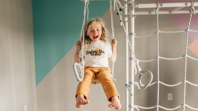 Laughing child riding a classic wood swing hanging from a Brainrich indoor play gym with rope net