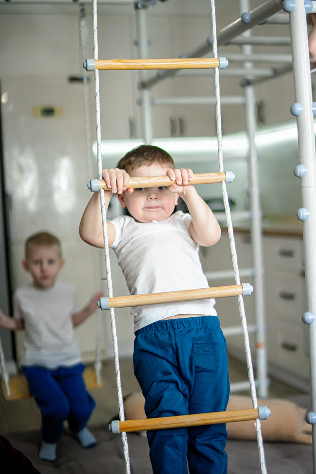 Preschooler climbing a rope ladder on a Brainrich steel play gym with his younger brother behind him