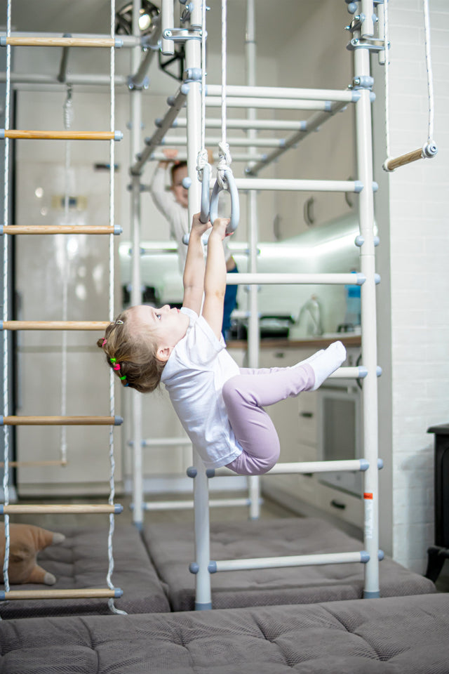 Toddler girl hanging from rings on a home play gym set up in a kitchen