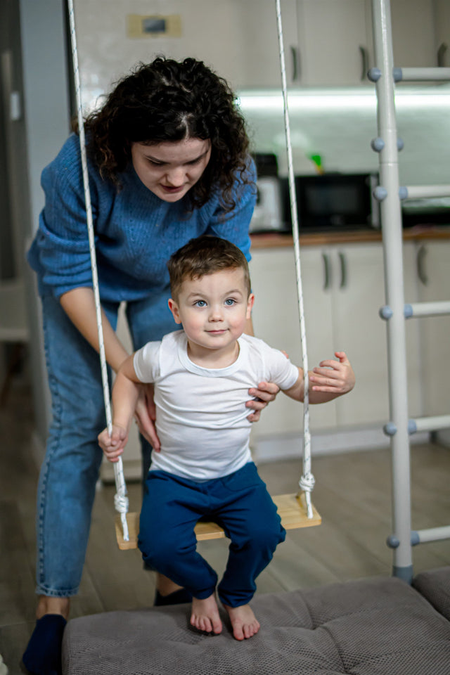 A mom standing behind her toddler on a wooden swing at home, part of a family's homeschool day