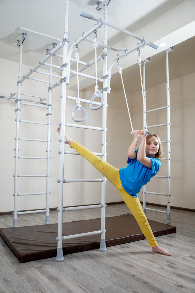 Girl holding the trapeze bar of a white Brainrich indoor play gym in a controlled stretch during a scheduled movement break at home