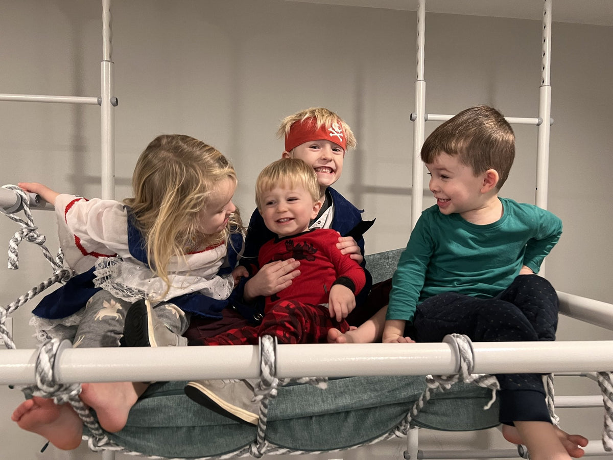Four kids laughing together in the net hammock of an indoor play gym in a real family playroom