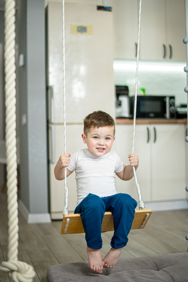 Toddler on a classic wooden sensory swing hanging from a Brainrich play gym frame with a mat below
