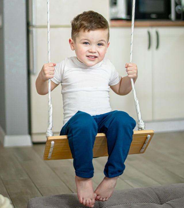 Boy sitting on a Brainrich Classic Swing wooden seat hung from an indoor steel play gym